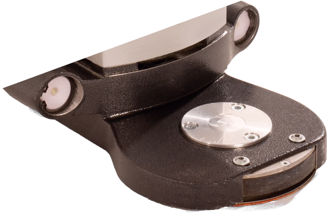 PALLMANN_dummy_Gecko Star attachment length short_msl_2018-07 Крупный план массивного литого металлического узла тёмно-коричневого/чёрного цвета с текстурированной поверхностью. Деталь имеет изогнутое основание и наклонный вверх фланец; по верхнему левому и правому краю видны два небольших круглых врезанных элемента, похожих на лампы или датчики. На плоской верхней поверхности установлена полированная круглая металлическая крышка с тремя видимыми винтами, окружённая несколькими потайными винтами. На переднем крае частично виден тонкий круглый вращающийся диск или тормозоподобный элемент с оранжевой кромкой. Изображение вырезано на белом фоне и демонстрирует детальную текстуру поверхности, головки винтов и металлические отражения.